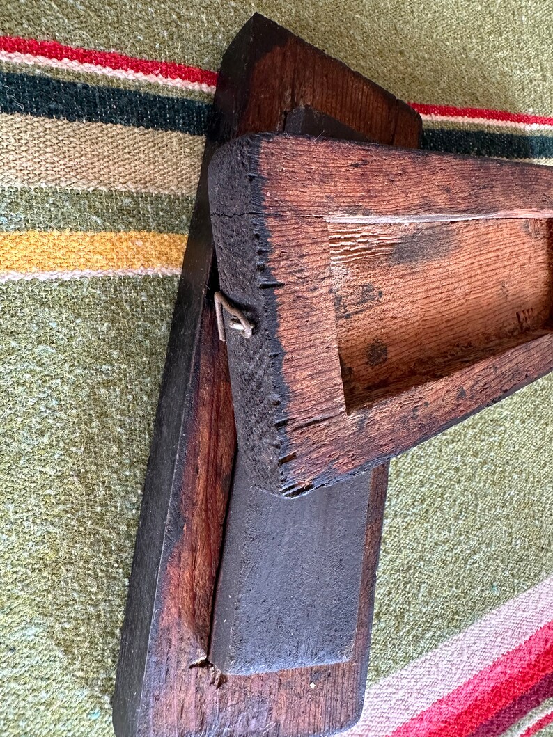 Op de afbeelding: Een verweerd, donkerbruin houten gereedschap met een rechthoekige basis en een verhoogd, open bovendeel. Het hout vertoont tekenen van ouderdom en slijtage, met zichtbare nerf en een metalen sluiting. Het gereedschap rust op een groen, rood en geel gestreept textiel.