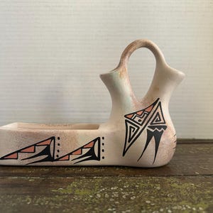 May include: A beige ceramic vessel with a rectangular base and a dual-spout top connected by a handle. The exterior features hand-painted black and salmon-colored geometric patterns, including stepped triangles and abstract linear designs.