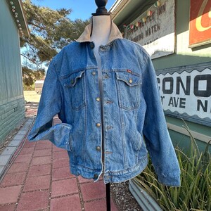 May include: A blue denim jacket featuring a tan corduroy collar, long sleeves, and a button-up front. The jacket includes two chest pockets with buttoned flaps and metal buttons throughout, displayed on a black mannequin torso.