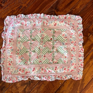 May include: A rectangular, floral-patterned pillow with a ruffled edge. The pillow features a patchwork design with red and green striped accents. The border is a red and white gingham check.