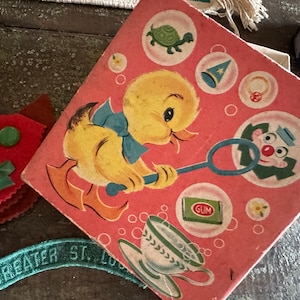 May include: A vintage pink box featuring a cartoon duck blowing bubbles, with a blue bow tie and orange feet. The box has illustrations of a turtle, a clown, a ring, and a teacup. A red felt cat and a green patch with "GREATER ST. LOUIS" are also visible.