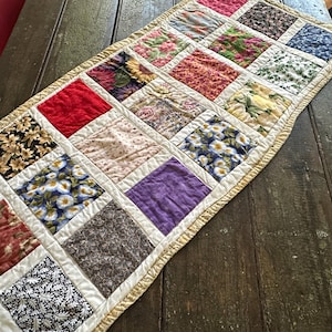 May include: A rectangular quilted table runner featuring a grid of assorted fabric squares in various colors and patterns, including floral, solid red, solid purple, and geometric prints. The runner is finished with a light yellow fabric border.