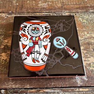 May include: A black decorative box with a Native American design. The box features a stylized figure in a colorful headdress and clothing, along with a magnifying glass design. The box is on a wooden surface.