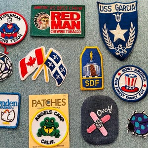 May include: A collection of embroidered patches with various designs. Includes a Superman Racing Team patch, a Red Man Chewing Tobacco patch, a USS Garcia patch, a soccer ball, Canadian and Quebec flags, and a patch with the text "Angels Camp Calif."