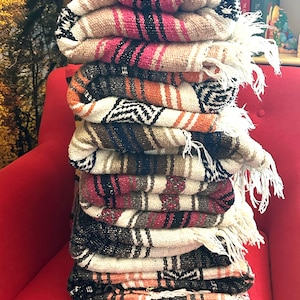 May include: A stack of seven folded woven blankets featuring various color combinations including pink, orange, brown, black, and white. Each blanket displays a repeating geometric pattern and includes white fringe detailing along the edges.