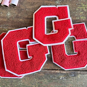 May include: Red chenille letter "G" patches with white borders. These embroidered patches are a classic design, perfect for adding to jackets, bags, or other fabric items. The patches are displayed on a rustic wooden surface.