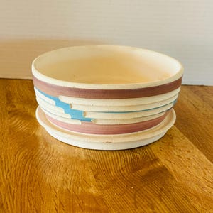 May include: A ceramic planter with a matching saucer. The planter is cream-colored with horizontal bands of maroon, light blue, and pink. The planter is round and has a wide opening. The saucer is also cream-colored.
