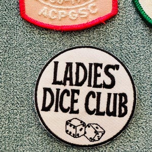Op de afbeelding: Een witte, ronde patch met de tekst "LADIES' DICE CLUB" in zwart, samen met twee dobbelstenen. De patch heeft een zwarte rand. Andere patches zijn zichtbaar op de achtergrond.
