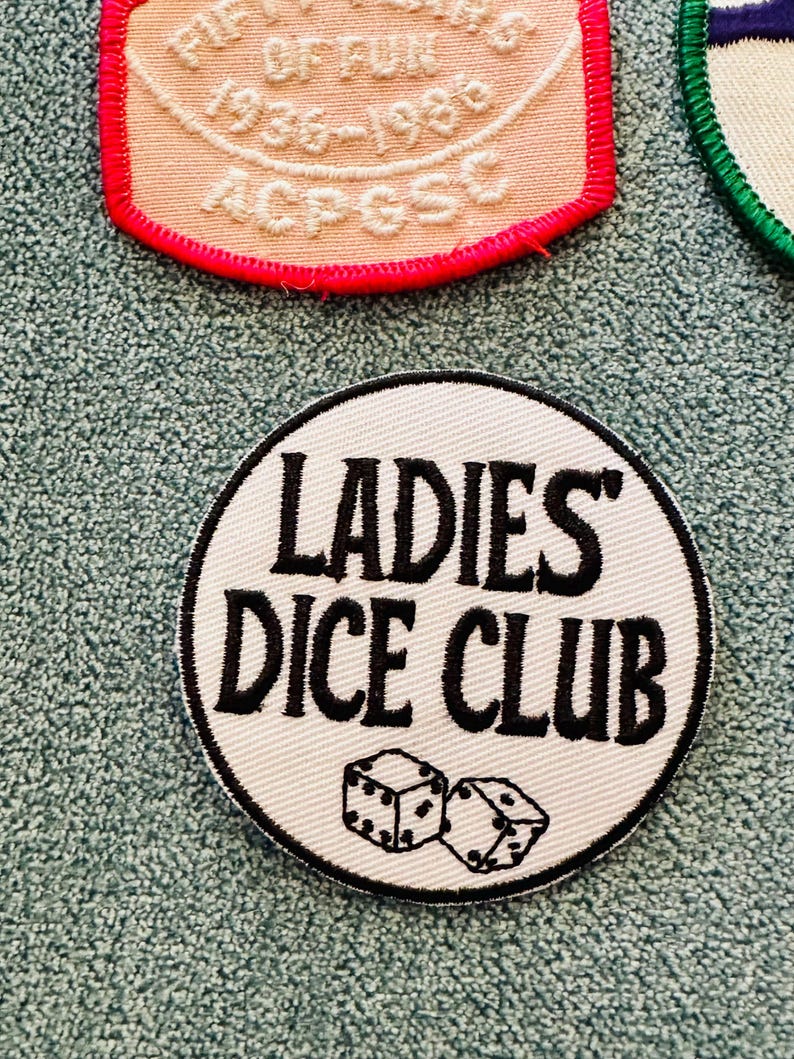 Op de afbeelding: Een witte, ronde patch met de tekst "LADIES' DICE CLUB" in zwart, samen met twee dobbelstenen. De patch heeft een zwarte rand. Andere patches zijn zichtbaar op de achtergrond.