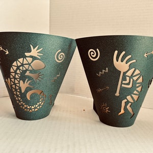 May include: Two dark teal metal cone-shaped candle holders. One features a lizard design, the other a Kokopelli figure, both in a lighter metallic color. Each has cut-out designs of arrows, spirals, and other shapes.