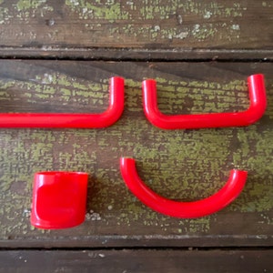 May include: A collection of bright red plastic hardware components, including two U-shaped handles, a curved piece, and a small rectangular piece. The items are arranged on a weathered, green-painted wooden surface.
