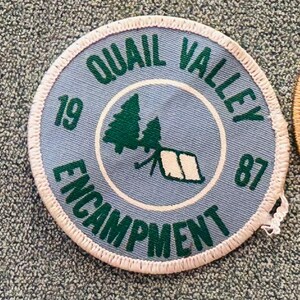Op de afbeelding: Een ronde, lichtblauwe patch met de woorden "Quail Valley Encampment" in groene letters. Het midden toont een groene boom- en tentafbeelding. De nummers "19" en "87" zijn ook aanwezig. De patch heeft een witte, gestikte rand.