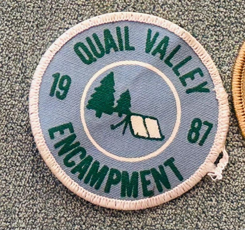 Op de afbeelding: Een ronde, lichtblauwe patch met de woorden "Quail Valley Encampment" in groene letters. Het midden toont een groene boom- en tentafbeelding. De nummers "19" en "87" zijn ook aanwezig. De patch heeft een witte, gestikte rand.