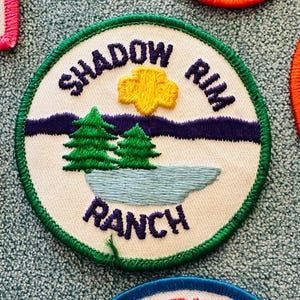 Op de afbeelding: Geborduurde patch met de woorden "Shadow Rim Ranch" in paarse letters. De patch heeft een groene rand, een gele zon, twee groene bomen en een blauw meer op een witte achtergrond. De patch is rond.