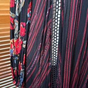 May include: A black garment with red stripes and floral patterned sleeves. The sleeves feature a black background with red, blue, and yellow flowers. The garment has a lace-like design down the front.