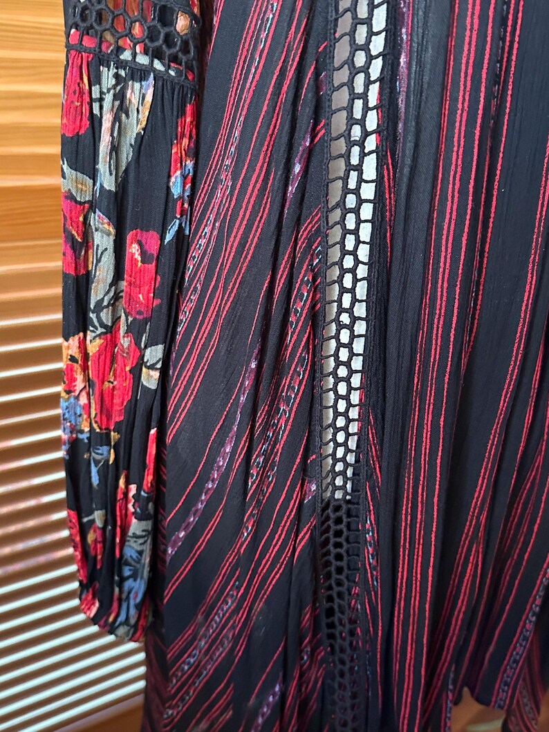 May include: A black garment with red stripes and floral patterned sleeves. The sleeves feature a black background with red, blue, and yellow flowers. The garment has a lace-like design down the front.