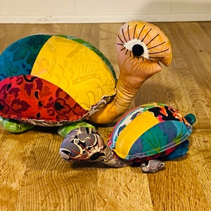 May include: Two plush turtle toys. The larger turtle has a shell made of colorful patchwork fabric in shades of blue, yellow, red, and green. The smaller turtle has a similar shell. Both have detailed faces and are on a wooden surface.
