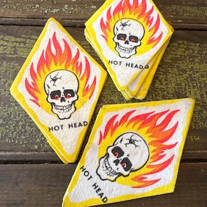 May include: A collection of yellow diamond-shaped patches with a skull design. The skull has red eyes and is surrounded by orange and red flames. The text "HOT HEAD" is printed below the skull.