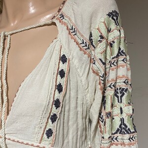 May include: Off-white long-sleeve top with intricate embroidery in shades of green, black, and peach. The top features a V-neckline with a drawstring and detailed patterns along the sleeves and bodice.