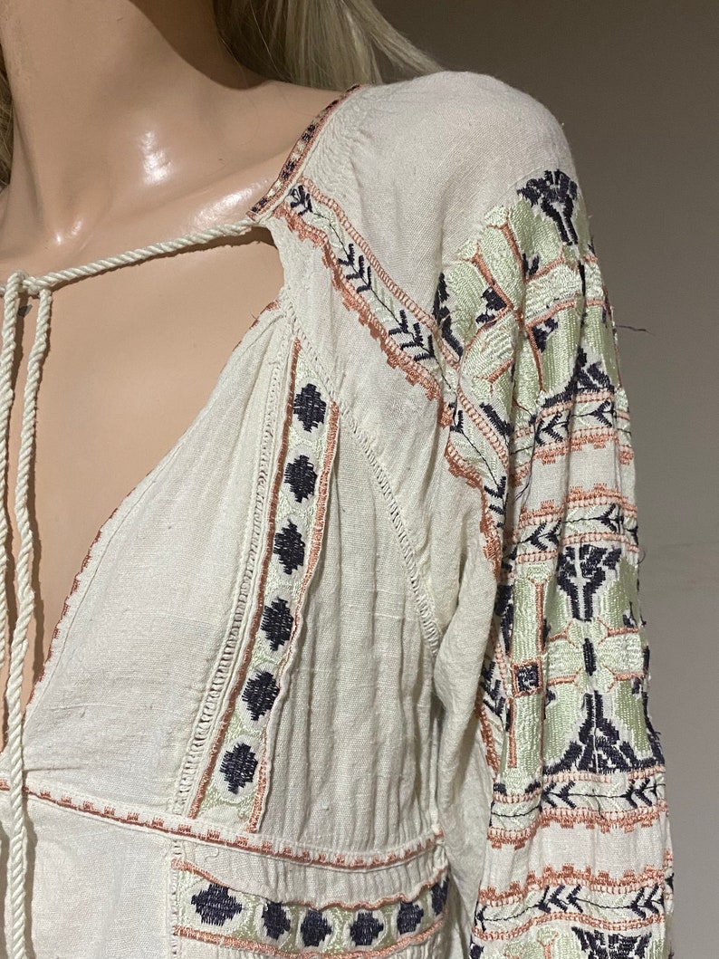 May include: Off-white long-sleeve top with intricate embroidery in shades of green, black, and peach. The top features a V-neckline with a drawstring and detailed patterns along the sleeves and bodice.