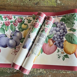 May include: Two rolls of wallpaper border featuring a colorful fruit design. The border has a cream background with illustrations of grapes, plums, pears, and other fruits. The top and bottom edges are a deep red.