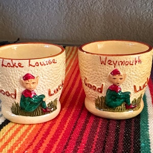 May include: Two vintage ceramic shot glasses. One reads "Lake Louise" and the other "Weymouth" in red script. Each features a seated elf figure in green and red, with "Good Luck" written on the side.