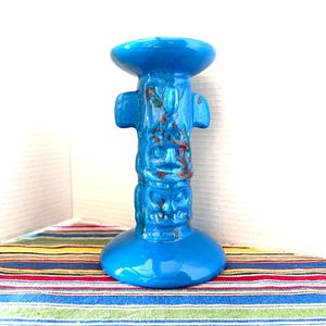 May include: A bright blue ceramic candle holder shaped like a totem pole. The cylindrical body features stacked, carved figures with subtle brown and green glaze accents. It has a wide, circular base and a flared top rim for holding a candle.
