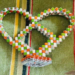 May include: A woven necklace with a heart-shaped design. The necklace is made of small, square, metallic, white, green, and orange pieces. The necklace is on a green and striped background.