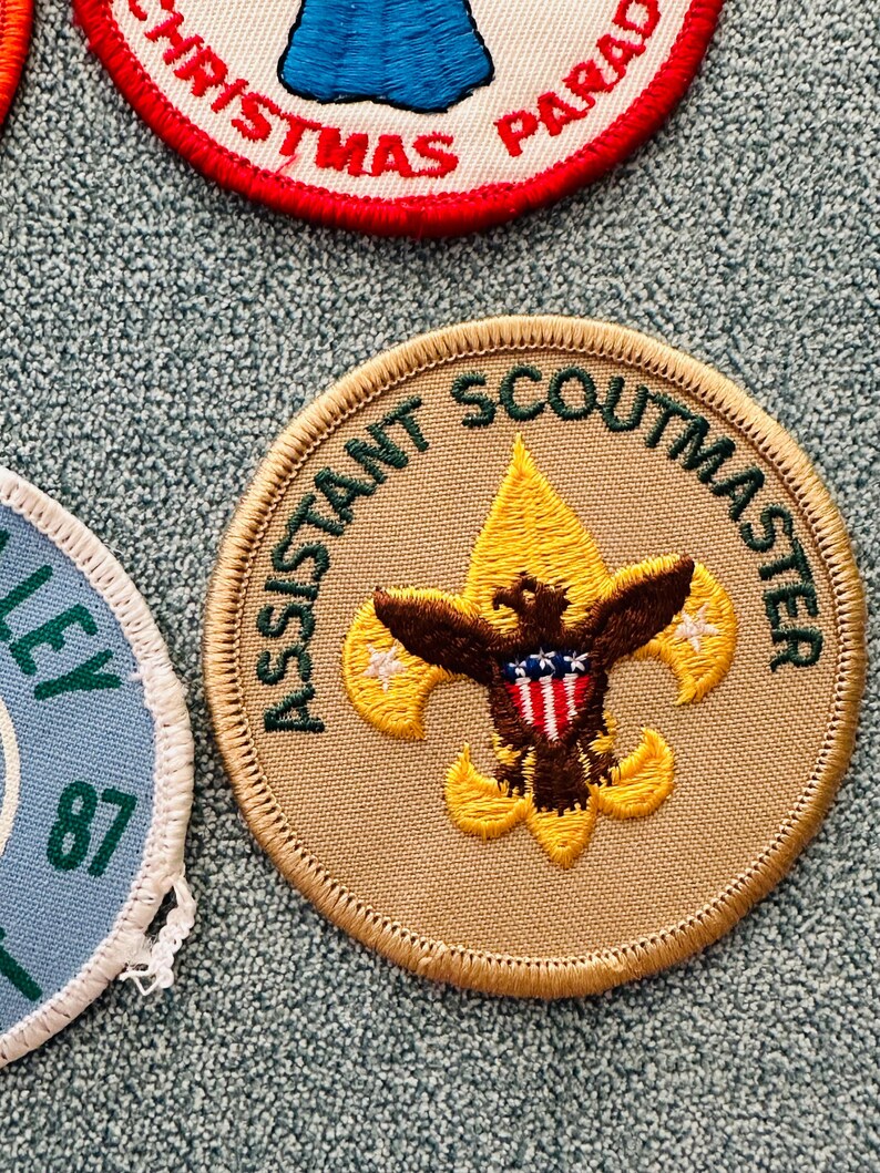 Op de afbeelding: Geborduurde Assistant Scoutmaster-patch met een beige achtergrond en gouden rand. De patch heeft een gouden fleur-de-lis met een bruine adelaar en een rood-wit-blauw schild. De tekst "ASSISTANT SCOUTMASTER" is groen.