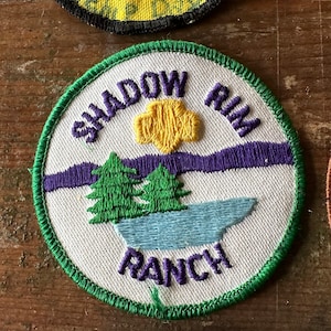 Op de afbeelding: Geborduurde ronde patch met een groene rand. Het ontwerp toont de woorden "SHADOW RIM RANCH" in paars en wit, een gele zon, paarse bergen, groene bomen en een blauw meer.