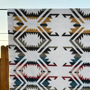 May include: White fabric with a geometric pattern in shades of brown, gold, teal, and red. The design features diamond and zigzag shapes. The fabric is hanging on a clothesline, with a wooden fence in the background.