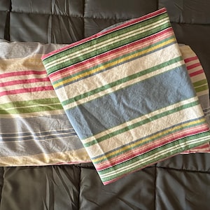 May include: Two striped pillowcases in the image. One pillowcase has stripes of pink, green, and yellow on a white background. The other pillowcase has stripes of blue, green, red, and yellow on a white background. The pillowcases are on a dark gray bedspread.