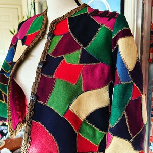 May include: A colorful patchwork jacket with a variety of fabric pieces in red, green, blue, and pink. The jacket has a brown ruffled trim and is displayed on a mannequin. The jacket is adorned with decorative buttons.