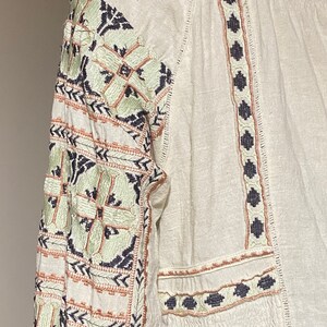May include: Off-white long-sleeve blouse with intricate embroidery. The sleeves and neckline feature detailed patterns in shades of green, navy, and peach. The blouse has a relaxed fit and a textured fabric.