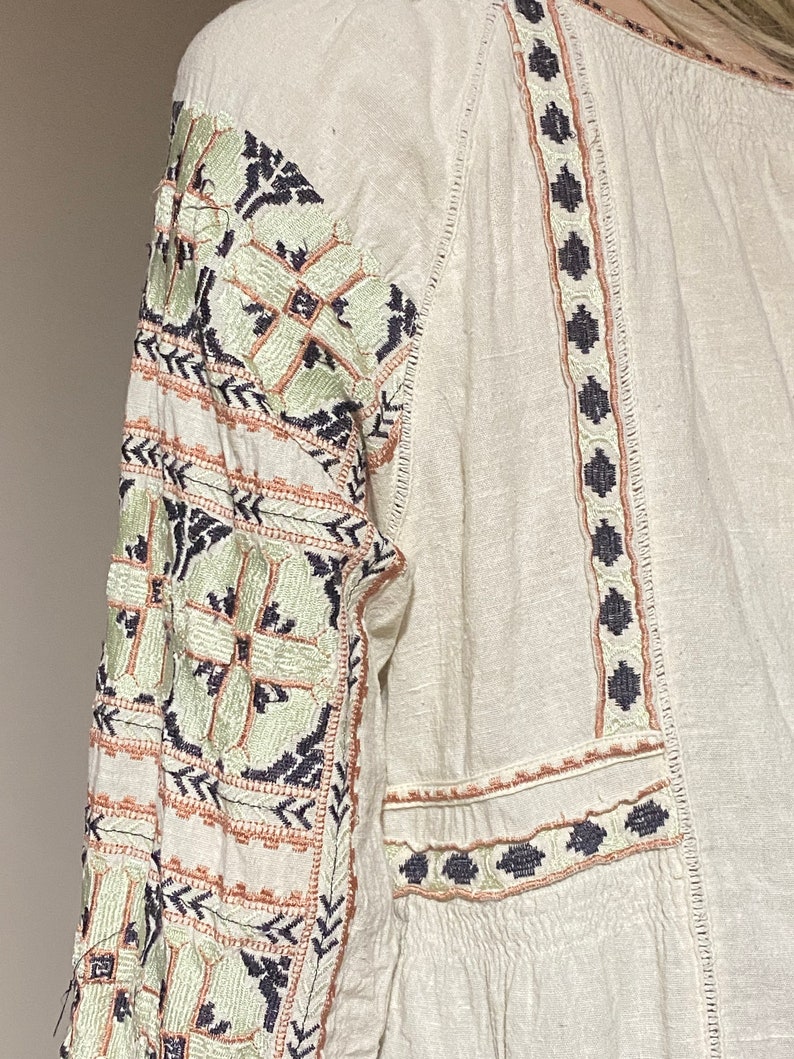 May include: Off-white long-sleeve blouse with intricate embroidery. The sleeves and neckline feature detailed patterns in shades of green, navy, and peach. The blouse has a relaxed fit and a textured fabric.