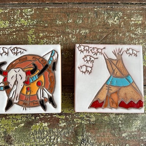 May include: Two square white ceramic tiles featuring hand-painted designs. The left tile displays a buffalo skull on a circular shield with feathers. The right tile depicts a conical tent with a herd of buffalo above. Both tiles use brown, red, and blue.