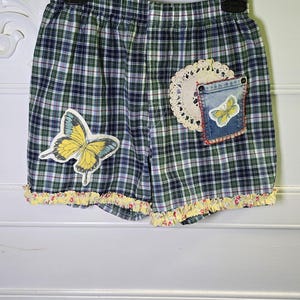 Puede incluir: Shorts a cuadros azules, verdes y blancos con una aplicación de mariposa amarilla. Los shorts tienen un ribete de volantes amarillos y un bolsillo de mezclilla con un parche de mariposa. Se adjunta un tapete de encaje blanco.