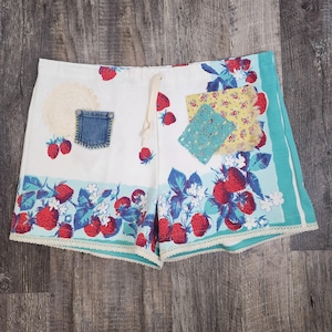 Puede incluir: Shorts de algodón blanco con estampado de fresas y ribete azul. Los shorts tienen cintura con cordón y están decorados con varios recortes de tela, incluyendo un parche de mezclilla, una servilleta de ganchillo y una tela floral amarilla y azul.