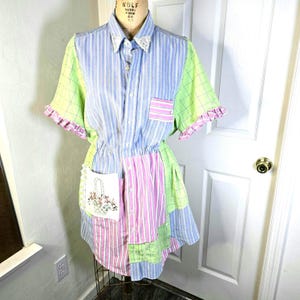 Peut inclure: Robe patchwork avec col et manches courtes. La robe présente un mélange de motifs rayés et à carreaux en bleu, rose et vert. Bords à volants et poche avec broderie florale.