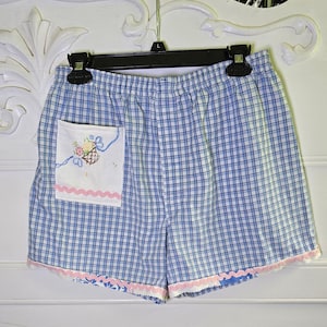 Puede incluir: Shorts a cuadros azules y blancos con un bolsillo blanco con un diseño de cesta floral bordada. Los shorts tienen un ribete festoneado rosa en el borde inferior. Los shorts están colgados de una percha negra.