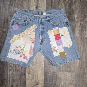 May include: Light blue denim shorts with patches of floral and patterned fabric. Patches include lace, floral embroidery, and colorful squares. The shorts have a button fly and frayed edges.