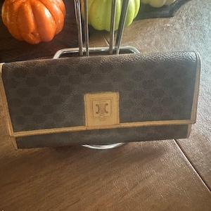 Puede incluir: Una cartera de cuero marrón con un patrón repetitivo y un ribete dorado. La cartera tiene forma rectangular y un emblema dorado en la parte delantera. La cartera está sobre una superficie de madera, con calabazas decorativas en el fondo.