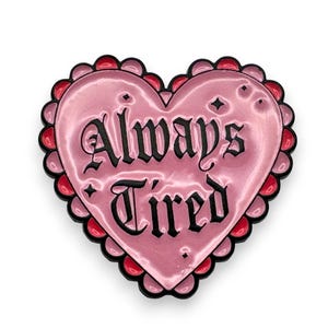 May include: A pink heart-shaped pin with the words "Always Tired" in a decorative font. The pin has a scalloped edge with red and black accents. The Riot Rhythm logo is in the bottom right corner.