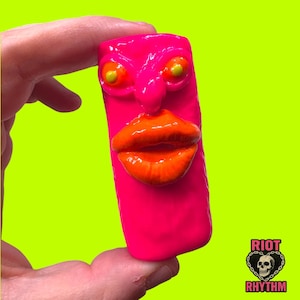 May include: A vibrant pink sculpture with a face featuring orange lips, a nose, and yellow-centered eyes. The sculpture is held in a hand against a bright green background. The text "RIOT RHYTHM" is visible.