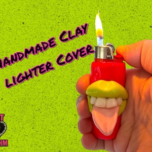 May include: A red lighter with a handmade clay cover shaped like a mouth with yellow lips, white teeth, and a pink tongue. The lighter is lit, with a flame. The background is bright green, with the text "HANDMADE CLAY LIGHTER COVER".