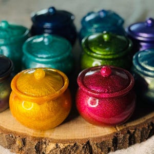 Puede incluir: Una colección de pequeños tarros redondos con tapa en varios colores, incluyendo verde azulado, verde, morado y dorado. Los tarros están colocados sobre una rodaja de madera, mostrando su acabado brillante e iridiscente. Cada tarro tiene un pequeño pomo en la tapa.