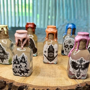 Puede incluir: Una colección de pequeñas botellas de vidrio transparente con etiquetas decorativas y sellos de cera de colores. Las botellas están dispuestas sobre una rodaja de madera, cada una con un diseño único, incluyendo un ojo, una calavera y una mano.