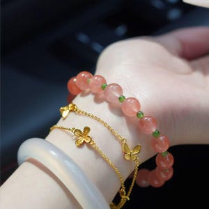 May include: A collection of bracelets featuring a white jade bangle, a gold chain bracelet with flower charms, and a gold chain bracelet with butterfly charms. A beaded bracelet with pink beads and green spacers is also visible.