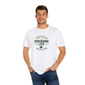 Puede incluir: Camiseta blanca con un gráfico verde que dice "PROPERTY OF YOUR NAME DEBUGGING LIFE ONE LINE AT A TIME." El diseño incluye un icono de error. La persona lleva vaqueros azules.
