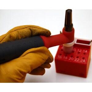 May include: A welding torch with a black handle and red nozzle is held by a person wearing yellow leather gloves. The torch is placed on a red block with numbered holes, likely for welding tips.